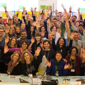 A large group of smiling workshop participants raising their hands together in a bright conference room